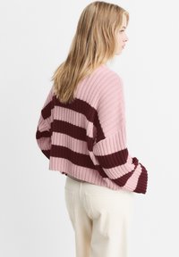 Blonde vrouw in een roze gebreide trui met donkerrode strepen, gecombineerd met hooglopende off-white broek, staande tegen een effen achtergrond.