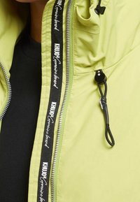 Limegrön jacka med dragkedjestängning, svart snörning och logotypdetalj, med en texturerad tyg och design med hög krage.