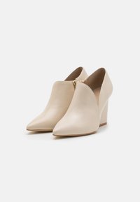 Para beżowych skórzanych botków za kostkę ze spiczastym noskiem, z bocznymi zamkami i blokowym obcasem na białym tle.