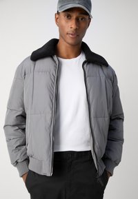 Veste rembourrée grise avec col en fausse fourrure noire. Elle dispose d'une fermeture éclair, de poignets élastiques et d'une coupe droite. Portée par-dessus une chemise blanche.