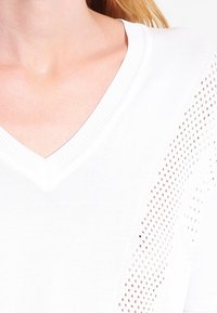 Weißes Strickoberteil mit V-Ausschnitt, seitlichen Mesh-Details, das ein strukturiertes Muster und atmungsaktiven Stoff betont.