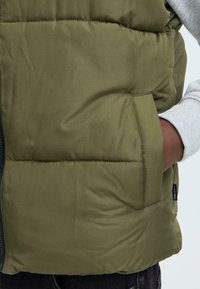 Olivgrüner Puffer-Weste mit Reißverschluss, gestepptem Muster, seitlicher Tasche und grau-gemusterten Sweaterärmeln, die darunter hervorlugen.