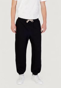 Zwarte sweatpants met een relaxed fit, een trekkoord in de taille en elastische boorden, gecombineerd met witte sneakers op een neutrale achtergrond.