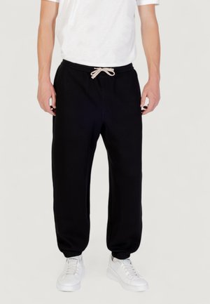 Zwarte sweatpants met een relaxed fit, een trekkoord in de taille en elastische boorden, gecombineerd met witte sneakers op een neutrale achtergrond.
