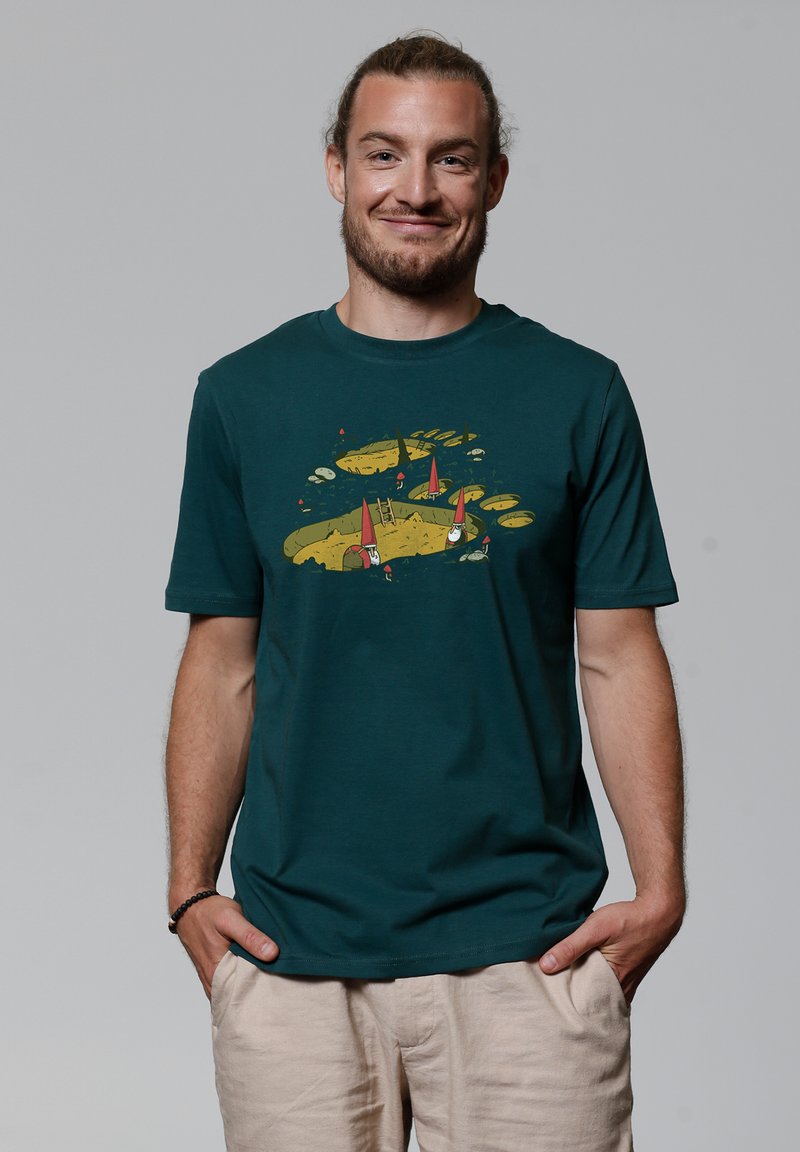 Donkergroene T-shirt met korte mouwen met een geel en rood grafisch ontwerp met gnoomachtige figuren en een ladder op een zandkleurige achtergrond.