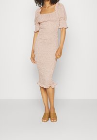 Robe cintrée jusqu'aux genoux en tissu rose doux avec un petit motif floral, décolleté carré, manches bouffantes et ourlet à volants. Associée avec des sandales tressées beiges.