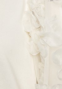 Haut blanc avec un tissu texturé et des volants floraux transparents d'un côté, mettant en valeur un design délicat et en relief.