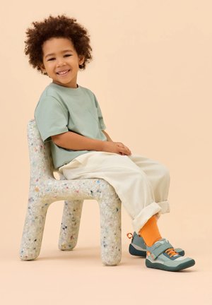 Kind mit lockigem Haar lächelt, während es auf einem weißen Stuhl aus recyceltem Kunststoff sitzt, trägt grünes Hemd, helle Hose, orangefarbene Socken und türkisfarbene Turnschuhe.