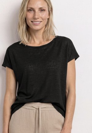 Blonde Frau trägt ein lockeres schwarzes Kurzarmtop und beige strukturierte Hosen mit einem gebundenen Taillenbund, steht vor einer schlichten hellen Wand.
