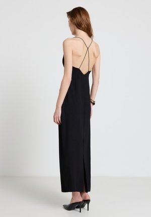 Donna che indossa un lungo abito nero senza schiena con sottili spalline incrociate e scarpe nere con tacchi alti, in piedi davanti a uno sfondo semplice.