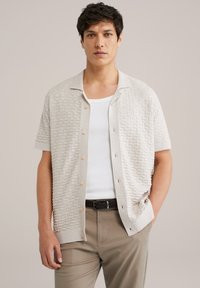 Maglietta a maniche corte di colore beige chiaro in maglia con un motivo testurizzato, bottoni in legno e colletto a camicia, abbinata a pantaloni marrone chiaro.