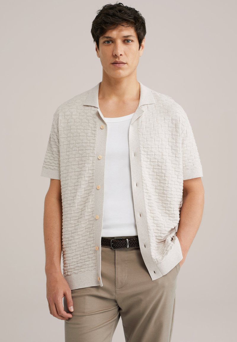 Maglietta a maniche corte di colore beige chiaro in maglia con un motivo testurizzato, bottoni in legno e colletto a camicia, abbinata a pantaloni marrone chiaro.
