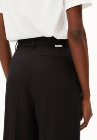Schwarze, weit geschnittene Hose aus glattem Material, mit einer Gesäßtasche und einem kleinen Etikett am Bund. Kombiniert mit einer locker sitzenden weißen Bluse.