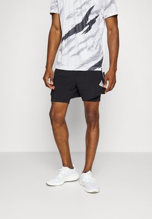 Mand iført sorte atletikshorts, en hvid og sort mønstret kortærmet skjorte og hvide løbesko, der står på en jævn gulvflade.