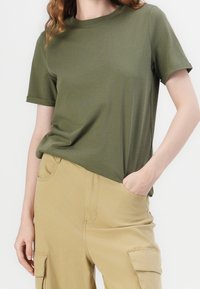 Kobieta w oliwkowej koszulce z krótkim rękawem, włożonej w beżowe spodnie cargo, z ręką w kieszeni, stojąca na tle białym.