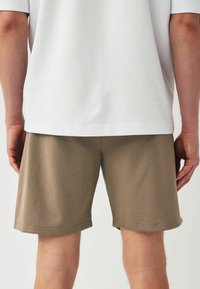 Shorts en coton marron avec un ourlet, vus de dos. Un t-shirt blanc partiellement visible, avec une coupe décontractée et des manches courtes.