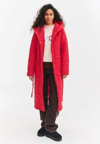Czerwona pikowana długa kurtka z kapturem, prezentowana na jasnym swetrze i ciemnych spodniach. Posiada przedni zamek błyskawiczny oraz teksturowaną tkaninę.