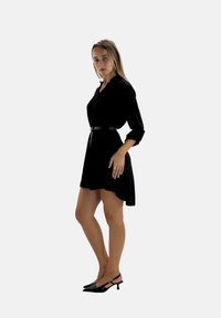 Zwarte overhemdjurk met kraag, lange mouwen, getailleerd met een dunne zwarte riem en een uitlopende zoom, gecombineerd met zwarte hakken.