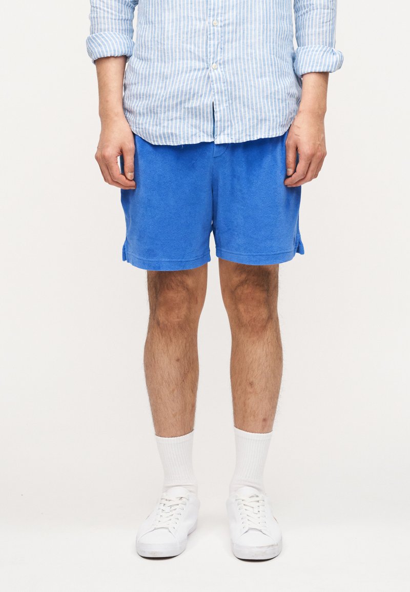 Άνδρας που φορά μπλε σορτς, λευκά α θλητικά παπούτσια και λευκές κάλτσες, όρθιος με τα χέρια χαλαρά δίπλα στο σώμα, μπροστά σε απλό φόντο.