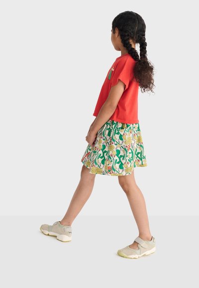 Fille aux cheveux tressés marchant, portant une chemise rouge, une jupe fleurie et des baskets beige sur un fond blanc uni.