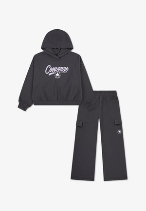 Juodas džemperis su kapišonu, ant kurio yra rožinės ir baltos spalvos „Converse“ logotipas, derinamas su juodomis cargo kelnėmis, kurios turi šonines kišenes ir logotipo detalę.