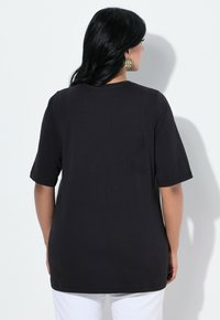 Una mujer con cabello negro largo lleva una camiseta negra holgada y grandes pendientes de aro dorados, de espaldas contra un fondo blanco.