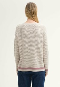 Beige gebreide trui met een visgraatpatroon, lange mouwen en een contrasterende bordeauxrode zoom. Van achter bekeken, gecombineerd met donkere blauwe jeans.