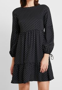 Vestido negro de lunares con mangas largas abullonadas, escote redondo y bajo con volantes. Presenta una tela suave y detalle de cordón en las muñecas.