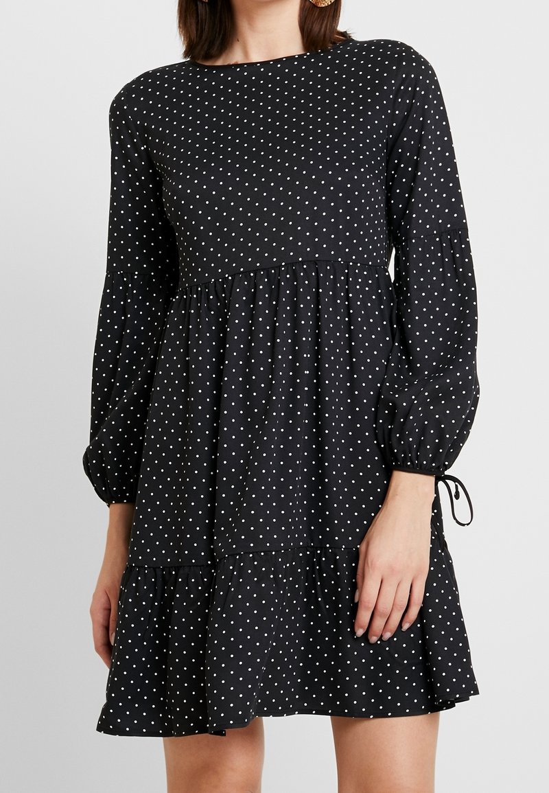 Vestido negro de lunares con mangas largas abullonadas, escote redondo y bajo con volantes. Presenta una tela suave y detalle de cordón en las muñecas.