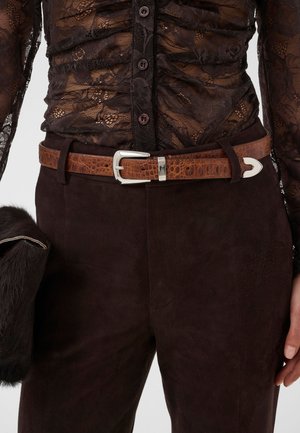 Ceinture en cuir marron texturé avec une boucle argentée portée par-dessus un pantalon marron foncé et un haut noir en dentelle transparente à boutons.