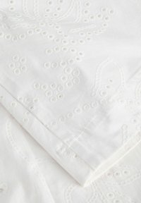 Tissu blanc avec une broderie ajourée détaillée présentant des motifs circulaires et floraux, plié pour montrer la texture et les coutures.