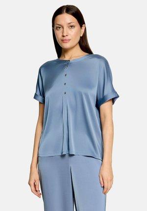 Femme portant un chemisier en satin bleu clair, ample, à manches courtes, avec des boutons sur le devant, et un pantalon large assorti.