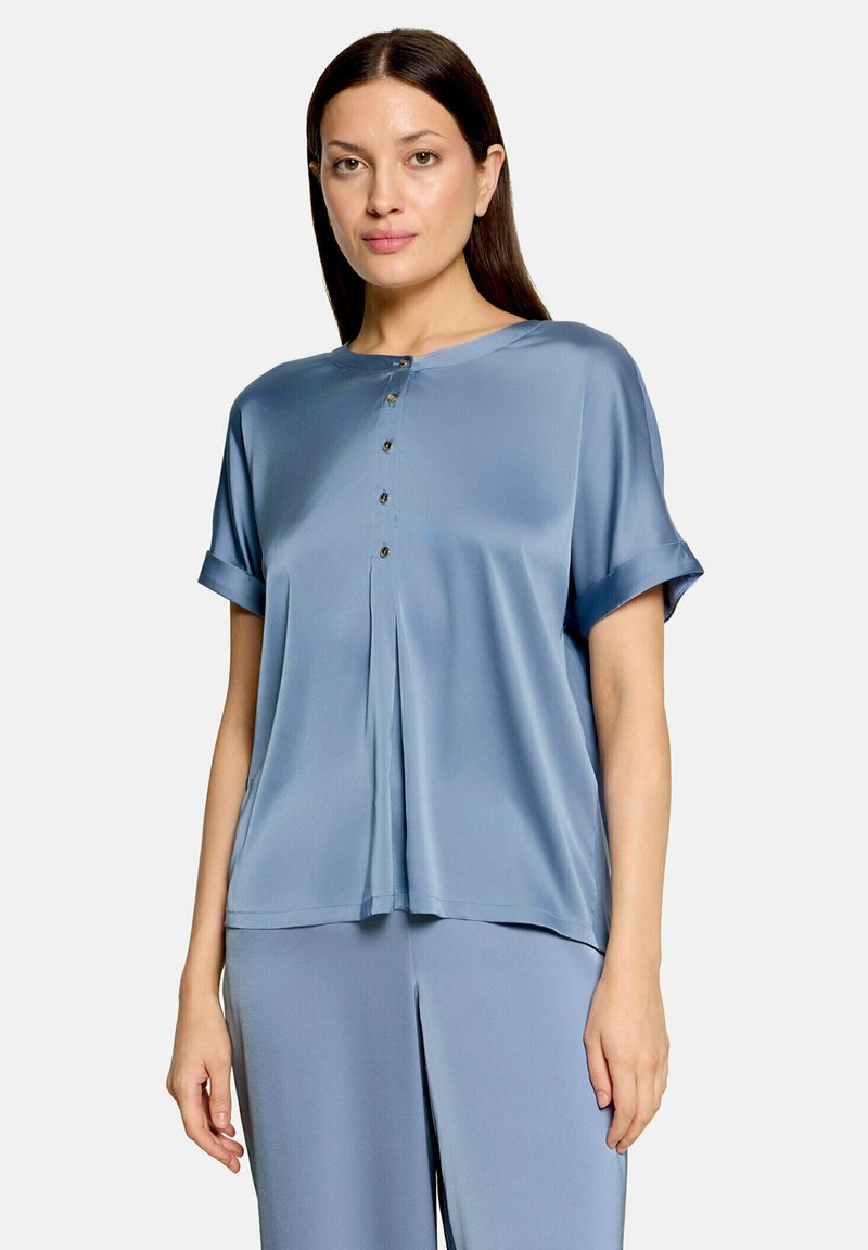 Femme portant un chemisier en satin bleu clair, ample, à manches courtes, avec des boutons sur le devant, et un pantalon large assorti.