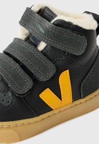 Musthave musta nahast beebi kinga kollase logoga, roheliste velcro rihmadega ja beeži fliisvoodriga beežil kummitaldal.