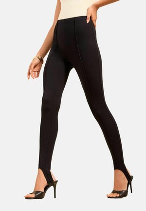 Kobieta ubrana w czarne legginsy z wysokim stanem i ozdobnym szwem z przodu oraz czarne sandały na wysokim obcasie, stoi z jedną ręką na biodrze.