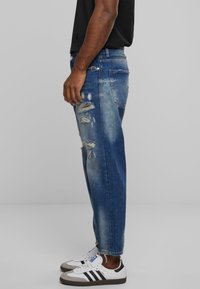 Vaqueros de mezclilla azul con detalles desgastados, desgastes ligeros y un diseño de pierna recta. Combinados con zapatillas blancas con rayas negras.