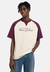 Κοντομάνικο t-shirt με εκρού σώμα και μανίκια raglan σε μαρόνο χρώμα. Διαθέτει σχεδιασμό με μοτίβο, κείμενο και λεπτομέρειες στο λαιμό με αντίθεση.