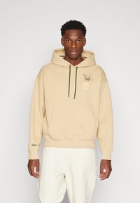 Bež dukserica s kapuljačom, prednjim džepom, zelenim vezicama i otisnutim tekstom u žutoj boji. Lacoste logo naglasak na lijevom rukavu. Mekana tekstura tkanine.