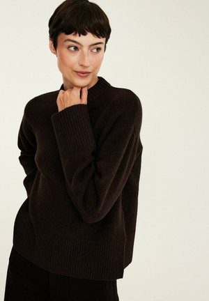 Femme aux cheveux courts et foncés portant un pull en maille brun foncé, regardant vers la gauche, la main droite touchant son menton sur un fond uni.
