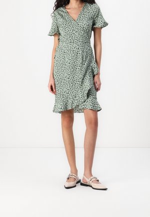 Vrouw draagt een groene wikkeljurk tot kniehoogte met zwarte en witte abstracte vlekken en nude platte schoenen met dubbele bandjes en gespen.