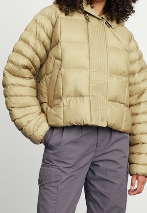 Personne portant une doudoune matelassée beige et un pantalon cargo gris, les mains dans les poches, montrée des épaules jusqu'aux hanches.