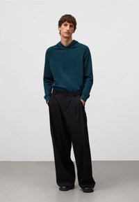 Felpa con cappuccio color teal dotata di lacci, abbinata a pantaloni neri plissettati. L'outfit presenta una vestibilità rilassata e una texture liscia, senza motivi visibili.