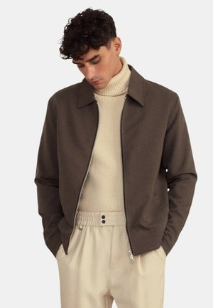 Homme aux cheveux bouclés et foncés portant une veste marron zippée par-dessus un pull à col roulé beige et un pantalon beige à taille élastique, les mains dans les poches.