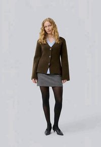 Kvinne med krøllete blondt hår, ikledd en brun knappet cardigan, blå skjorte, svart og hvit rutet skjørt, svarte strømpebukser og svarte hæler.