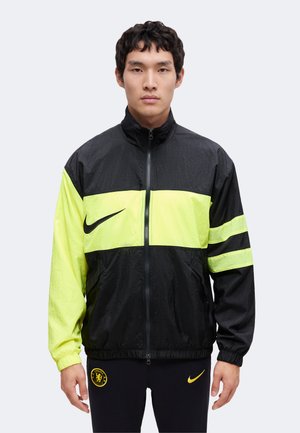 Junger Mann trägt eine schwarz-neongelbe Reißverschlussjacke mit Nike-Logo und steht vor einem schlichten hellen Hintergrund.