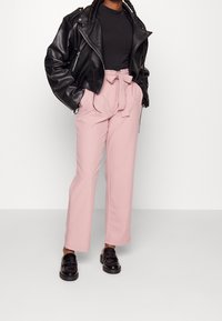 Zwarte leren motorjack over een zwart shirt, gecombineerd met hoog uitgesneden roze broek met een strik in de taille en zwarte instappers.