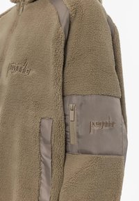Giacca in pile beige con tessuto testurizzato, tasca laterale con zip e pannelli delle maniche in tessuto liscio a contrasto. Presenta un logo ricamato sul petto e sulla manica.