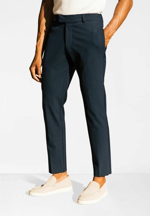 Granatowe, dopasowane spodnie o przednich zakładkach, z bocznymi kieszeniami i wąskim krojem, zestawione z beżowymi butami wsuwanymi o fakturowanej powierzchni.