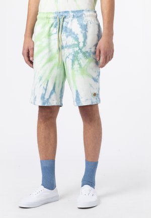 Mann trägt grün-blaue Batik-Shorts, blaue Crew-Socken und weiße Low-Top-Sneakers, steht vor weißem Hintergrund.