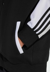 Crna dukserica s bijelim i crnim prugastim rukavima i bočnim džepovima sa patentnim zatvaračem, izrađena od mekane teksturirane tkanine.